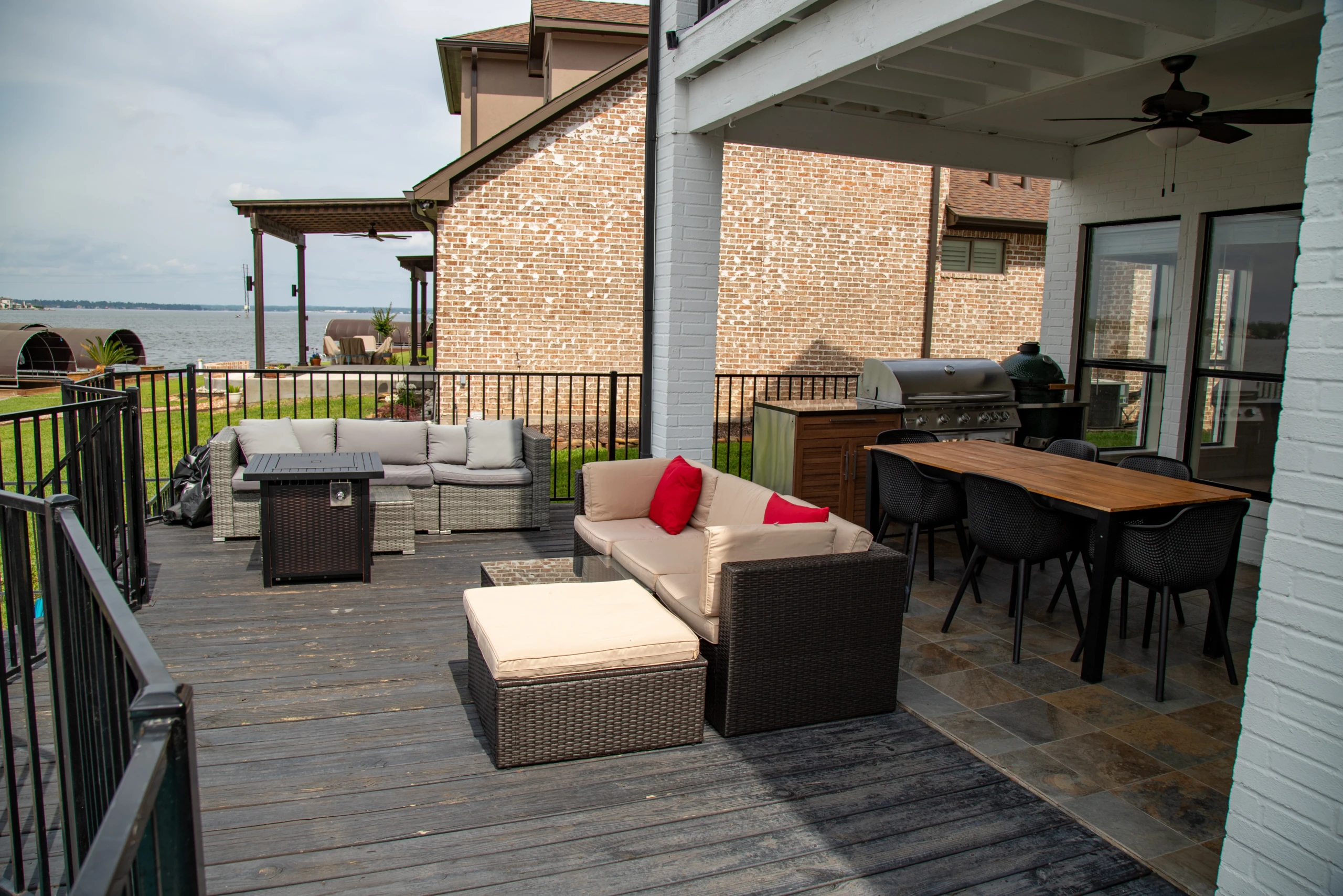 Covered lakeside patio with wicker lounge seating, accent pillows, and a gas grill overlooking calm water, ideal for cooler