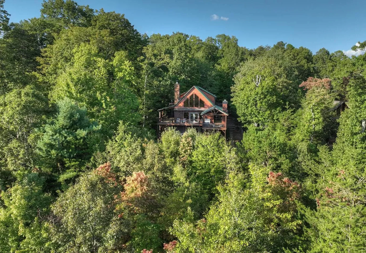 Modern chalet-style home with A-frame gables and a wooden deck in a dense forest, a Hot tub cabin deck for a Blue Ridge