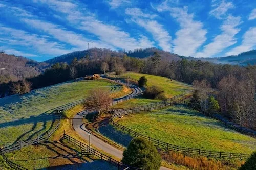 Aerial view of rolling green pastures and rustic wooden fencing below forested mountains in a Blue Ridge GA rural property,