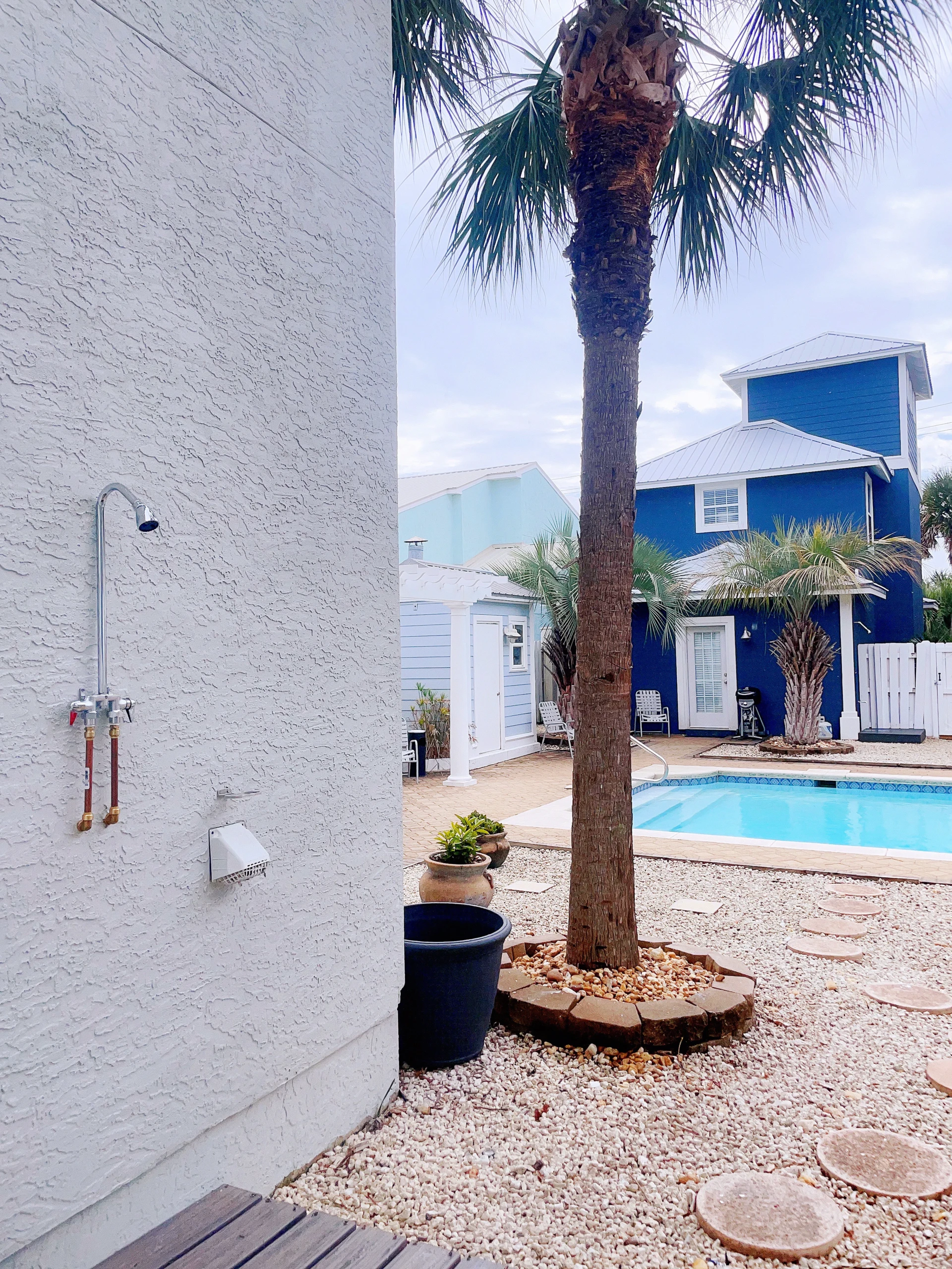 Backyard with bright blue pool, white textured wall with outdoor shower, and palm trees at Sea La Vie, a Panama City Beach,