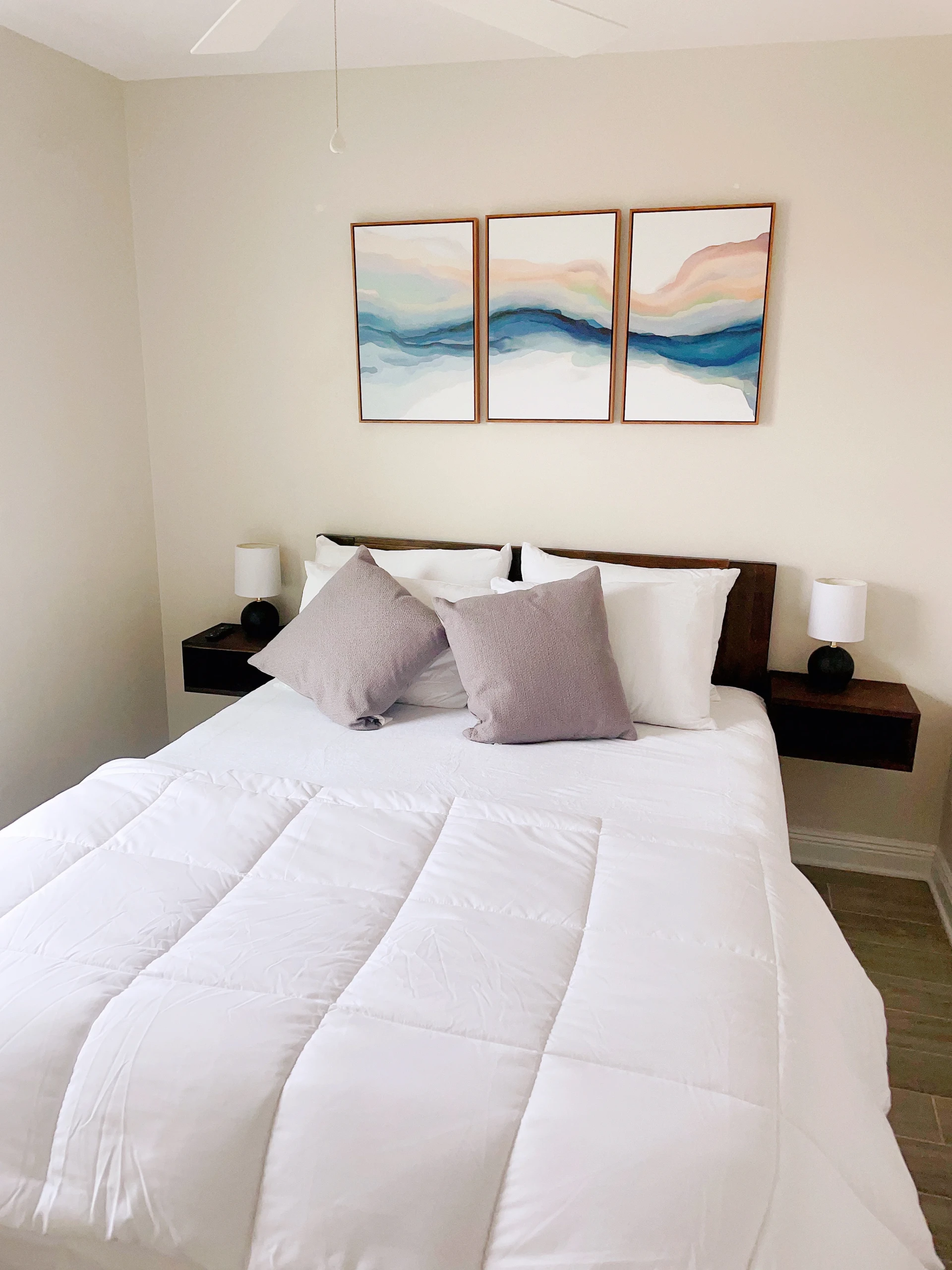 Modern bedroom with white queen bed, gray pillows, dark nightstands, and blue-peach watercolor triptych art, Sea La Vie